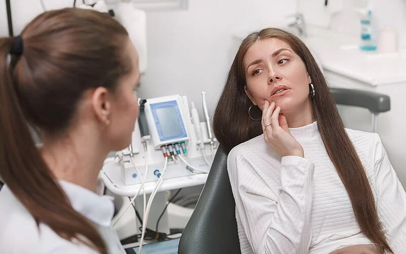 Eine Frau sitzt auf dem Behandlungsstuhl in einer zahnärztlichen Praxis. Sie fasst sich mit der Hand an den rechten Unterkiefer. Ihr Gegenüber ist eine Zahnärztin, die der Patientin zuhört.