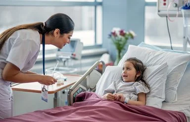 Ein kleines Mädchen liegt in einem Krankenbett, eine Kinderpflegefachperson beugt sich zu dem Mädchen herunter. Beide lachen sich an.