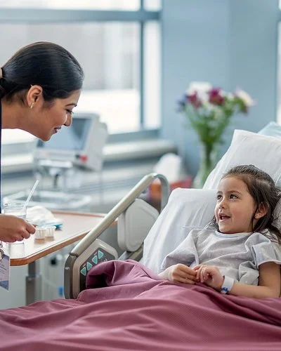 Ein kleines Mädchen liegt in einem Krankenbett, eine Kinderpflegefachperson beugt sich zu dem Mädchen herunter. Beide lachen sich an.