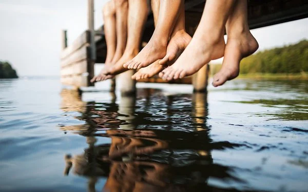 Vier Menschen sitzen auf einem Bootssteg an einem Gewässer. Zu sehen sind ihre nackten Unterschenkel und die Füße.