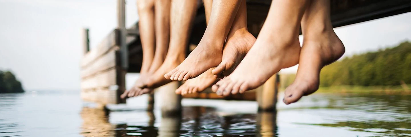 Vier Menschen sitzen auf einem Bootssteg an einem Gewässer. Zu sehen sind ihre nackten Unterschenkel und die Füße.
