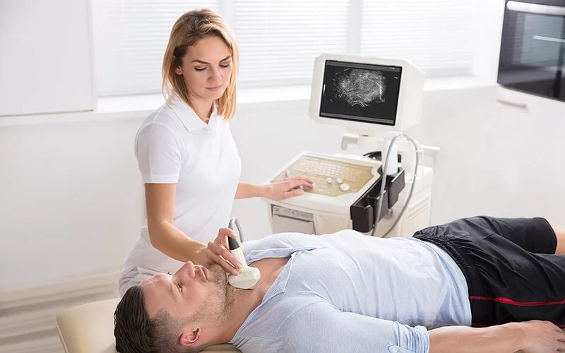 Ein Mann liegt auf dem Rücken auf einer Liege in einer ärztlichen Praxis. Eine Ärztin untersucht seinen Hals mit einem Ultraschallgerät.