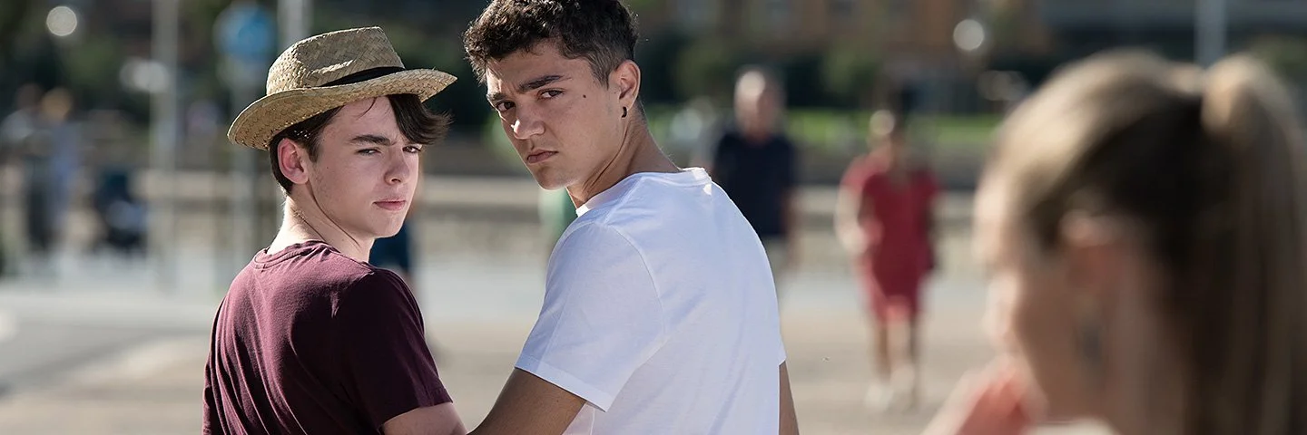 Zwei junge Männer im öffentlichen Raum umarmen sich von hinten, drehen sich um und schauen ernst, weil eine rechts im Bild unscharf zu sehende Frau ihnen etwas zuruft.