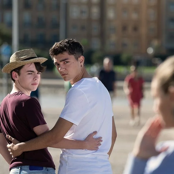 Zwei junge Männer im öffentlichen Raum umarmen sich von hinten, drehen sich um und schauen ernst, weil eine rechts im Bild unscharf zu sehende Frau ihnen etwas zuruft.