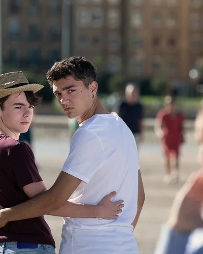 Zwei junge Männer im öffentlichen Raum umarmen sich von hinten, drehen sich um und schauen ernst, weil eine rechts im Bild unscharf zu sehende Frau ihnen etwas zuruft.