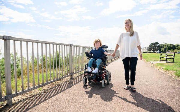 Mutter geht Hand in Hand mit ihrem Kind, das wegen einer Behinderung im Rollstuhl sitzt, spazieren.