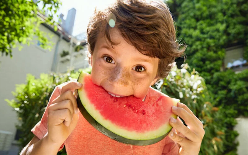 Kind schaut in die Kamera und beißt in eine Wassermelone rein.