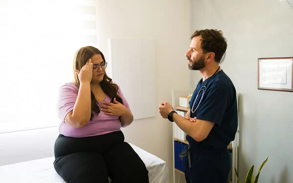 Eine Patientin mit Übergewicht sitzt im Behandlungszimmer auf einer Arztliege und spricht mit dem Arzt.