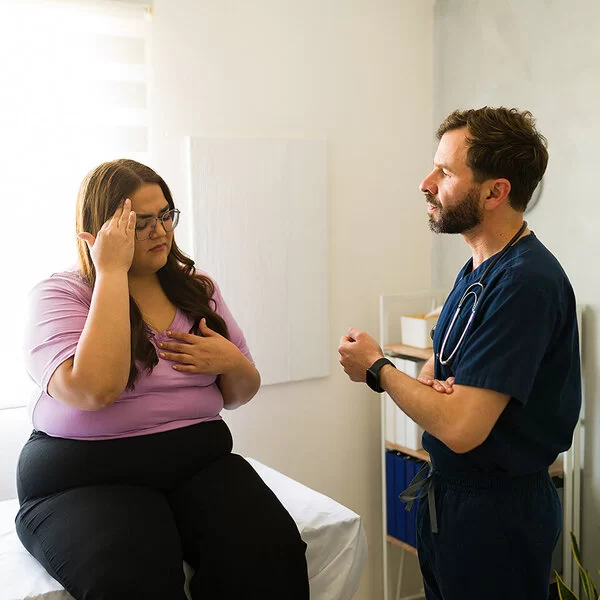 Eine Patientin mit Übergewicht sitzt im Behandlungszimmer auf einer Arztliege und spricht mit dem Arzt.