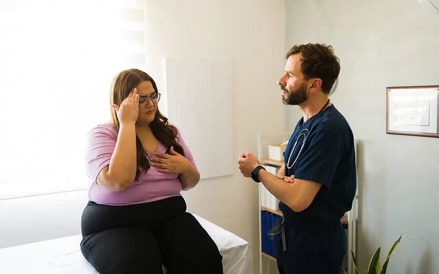 Eine Patientin mit Übergewicht sitzt im Behandlungszimmer auf einer Arztliege und spricht mit dem Arzt.