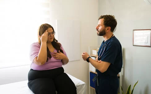 Eine Patientin mit Übergewicht sitzt im Behandlungszimmer auf einer Arztliege und spricht mit dem Arzt.