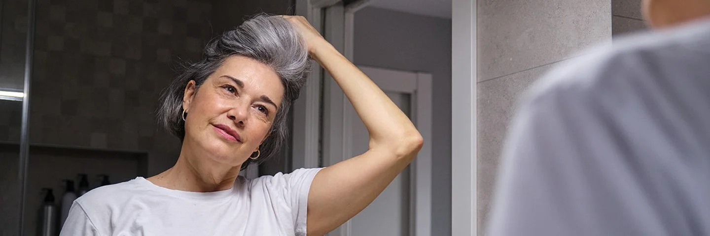Eine Frau betrachtet sich im Spiegel und fasst sich in ihr graues Haar.