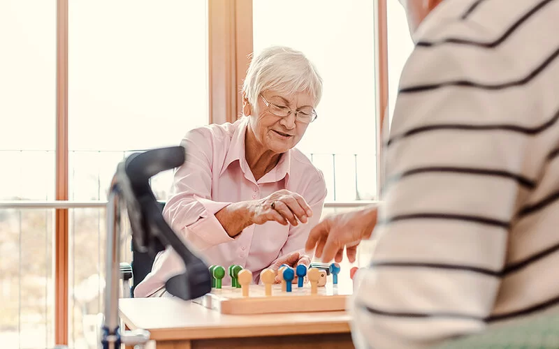 Man sieht zwei ältere Menschen, die an einem Tisch sitzen und spielen. Auf dem Tisch eine hölzerne Spielunterlage mit großen, farbigen Spielfiguren aus Holz, die in das Spielbrett gesteckt werden.