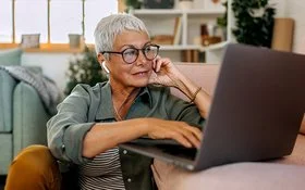 Eine Frau mit kurzen weißen Haaren und großer Brille sitzt in lockerer Kleidung auf dem Boden ihres Wohnzimmers vor dem Sofa. Auf dem Sofa steht ein geöffnetes Notebook, die Frau schaut auf das Display und bedient das Touchpad mit ihrer rechten Hand.