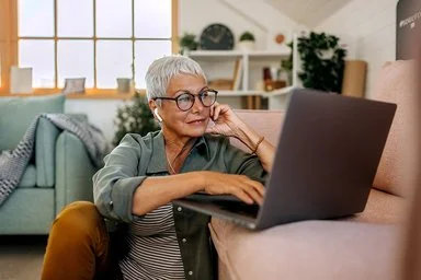 Eine Frau mit kurzen weißen Haaren und großer Brille sitzt in lockerer Kleidung auf dem Boden ihres Wohnzimmers vor dem Sofa. Auf dem Sofa steht ein geöffnetes Notebook, die Frau schaut auf das Display und bedient das Touchpad mit ihrer rechten Hand.