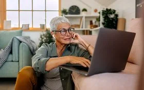 Eine Frau mit kurzen weißen Haaren und großer Brille sitzt in lockerer Kleidung auf dem Boden ihres Wohnzimmers vor dem Sofa. Auf dem Sofa steht ein geöffnetes Notebook, die Frau schaut auf das Display und bedient das Touchpad mit ihrer rechten Hand.