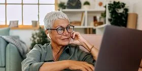 Eine Frau mit kurzen weißen Haaren und großer Brille sitzt in lockerer Kleidung auf dem Boden ihres Wohnzimmers vor dem Sofa. Auf dem Sofa steht ein geöffnetes Notebook, die Frau schaut auf das Display und bedient das Touchpad mit ihrer rechten Hand.