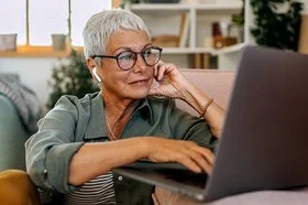 Eine Frau mit kurzen weißen Haaren und großer Brille sitzt in lockerer Kleidung auf dem Boden ihres Wohnzimmers vor dem Sofa. Auf dem Sofa steht ein geöffnetes Notebook, die Frau schaut auf das Display und bedient das Touchpad mit ihrer rechten Hand.