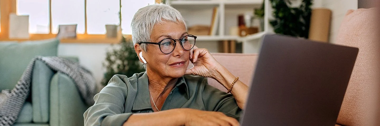 Eine Frau mit kurzen weißen Haaren und großer Brille sitzt in lockerer Kleidung auf dem Boden ihres Wohnzimmers vor dem Sofa. Auf dem Sofa steht ein geöffnetes Notebook, die Frau schaut auf das Display und bedient das Touchpad mit ihrer rechten Hand.