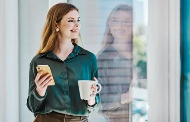 Eine junge Frau blickt lächelnd auf ihr Spiegelbild im Fenster. In der einen Hand hält sie eine Tasse, in der anderen ein Handy.