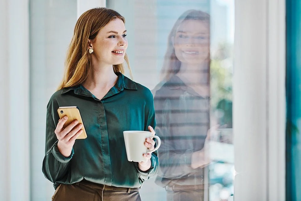 Eine junge Frau blickt lächelnd auf ihr Spiegelbild im Fenster. In der einen Hand hält sie eine Tasse, in der anderen ein Handy.