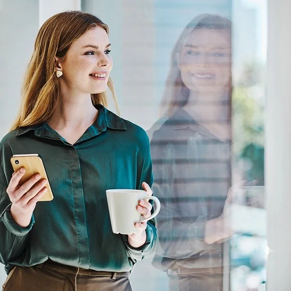 Eine junge Frau blickt lächelnd auf ihr Spiegelbild im Fenster. In der einen Hand hält sie eine Tasse, in der anderen ein Handy.