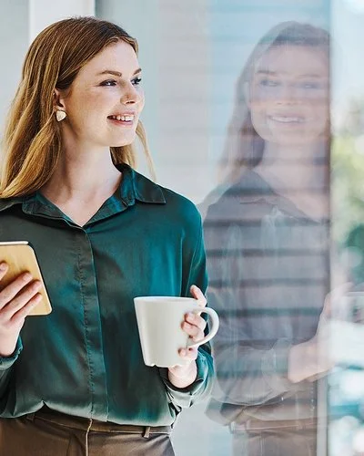 Eine junge Frau blickt lächelnd auf ihr Spiegelbild im Fenster. In der einen Hand hält sie eine Tasse, in der anderen ein Handy.