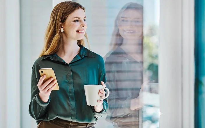 Eine junge Frau blickt lächelnd auf ihr Spiegelbild im Fenster. In der einen Hand hält sie eine Tasse, in der anderen ein Handy.