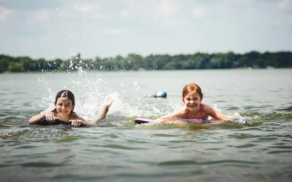 Zwei Kinder auf Bodyboards paddeln lachend im flachen Wasser eines Sees und spritzen dabei Wasser hoch.