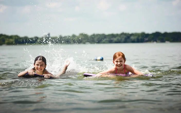 Zwei Kinder auf Bodyboards paddeln lachend im flachen Wasser eines Sees und spritzen dabei Wasser hoch.