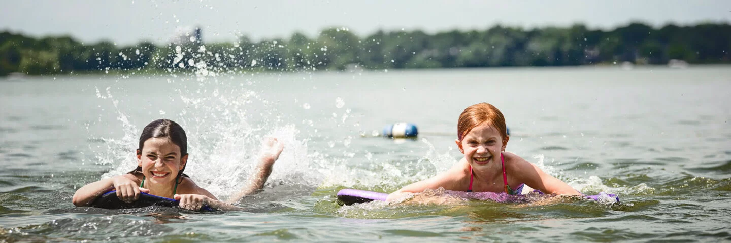 Zwei Kinder auf Bodyboards paddeln lachend im flachen Wasser eines Sees und spritzen dabei Wasser hoch.