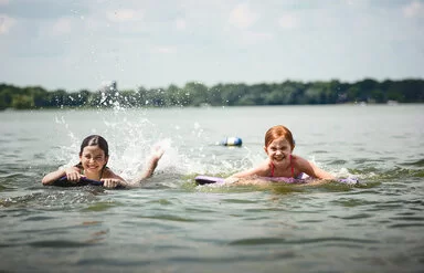 Zwei Kinder auf Bodyboards paddeln lachend im flachen Wasser eines Sees und spritzen dabei Wasser hoch.