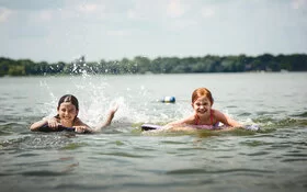 Zwei Kinder auf Bodyboards paddeln lachend im flachen Wasser eines Sees und spritzen dabei Wasser hoch.