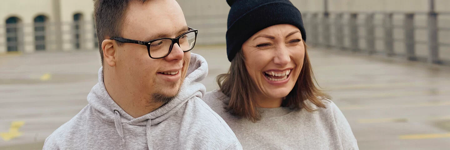 Ein Mann und eine Frau stehen in grauen Pullis auf der Straße und lachen. 