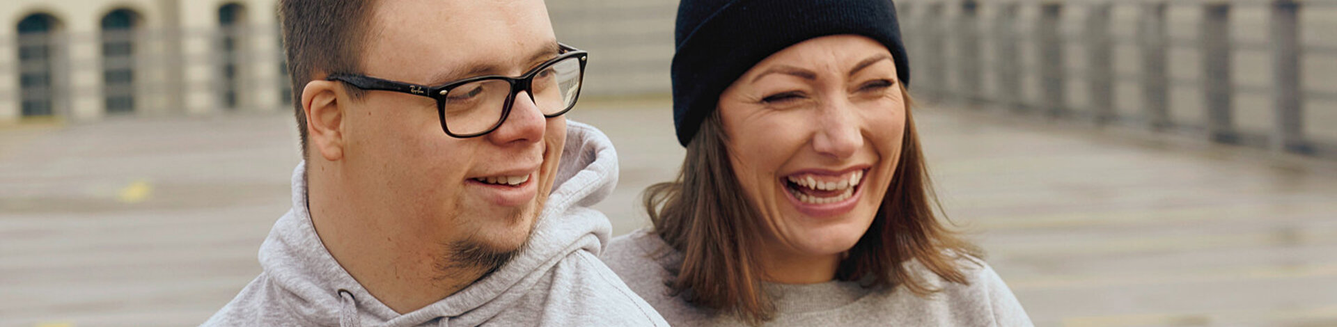 Ein Mann und eine Frau stehen in grauen Pullis auf der Straße und lachen. 