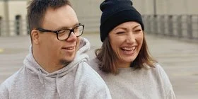 Ein Mann und eine Frau stehen in grauen Pullis auf der Straße und lachen.