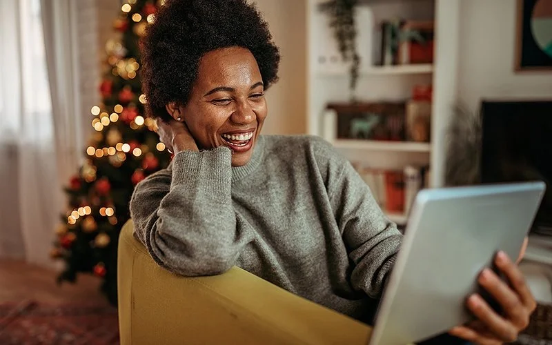 Eine Frau sitzt im weihnachtlich geschmückten Wohnzimmer, sie lacht und macht einen Video-Call über ihr Tablet.