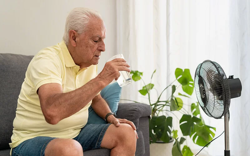 Ein älterer Mann sitzt bei geschlossenen Vorhängen auf einem Sofa in seinem Wohnzimmer vor einem Ventilator und trinkt ein Glas Wasser.
