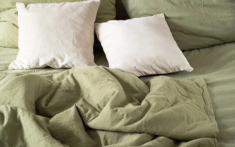 Ein von vorn und leicht oben fotografiertes Bett, das Bild rundherum stark angeschnitten: zwei mit salbeigrüner Leinenbettwäsche bezogene Kopfkissen und zwei Bettdecken. Auf den Kopfkissen liegen zwei kleinere naturweiße Kissen. Eine der Decken liegt zerwühlt im Vordergrund.