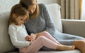 Kleines Mädchen sitzt neben seiner Mutter auf der Couch und hält sich den Bauch.