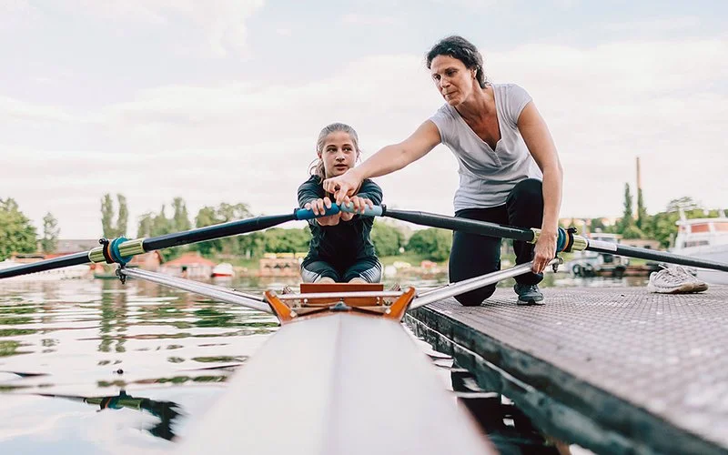 Ein Mädchen in einem Ruderboot. Eine Frau zeigt ihr, wie sie die Ruder hält.