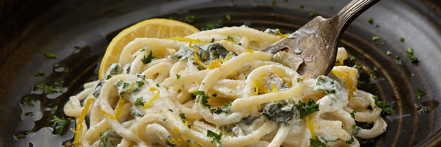 Ein dunkler Keramikteller, darauf Spaghetti mit Spinat und Ricotta, garniert mit einem Stück Zitrone.