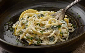 Ein dunkler Keramikteller, darauf Spaghetti mit Spinat und Ricotta, garniert mit einem Stück Zitrone.