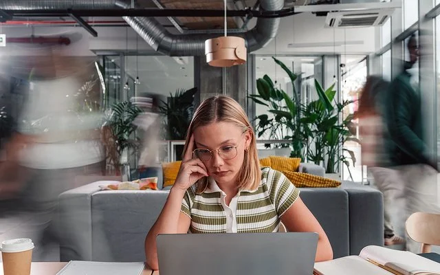 Eine Frau sitzt in einem Büro und blickt angestrengt auf einen Laptop, um sie herum hektische Kollegen und Kolleginnen.