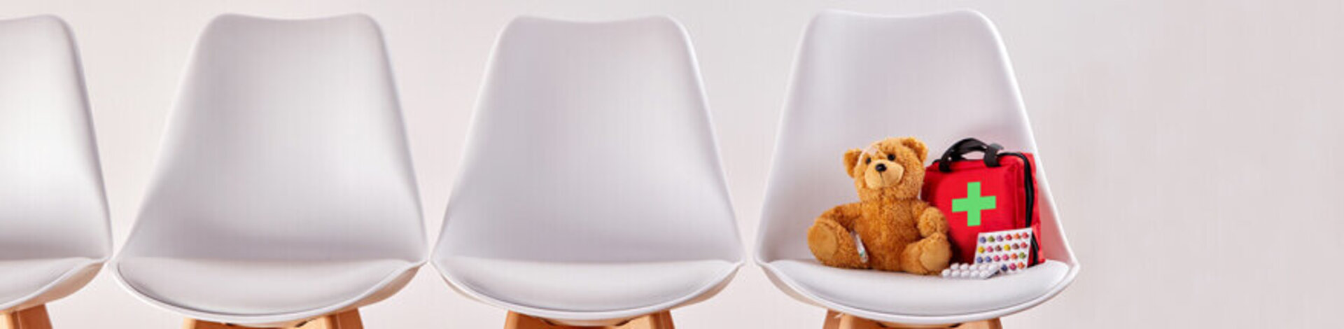 Das Bild zeigt vier weiße nebeneinander stehende Stühle. Auf dem Stuhl ganz rechts befindet sich ein Teddybär und ein kleiner Erste-Hilfe-Rucksack. 