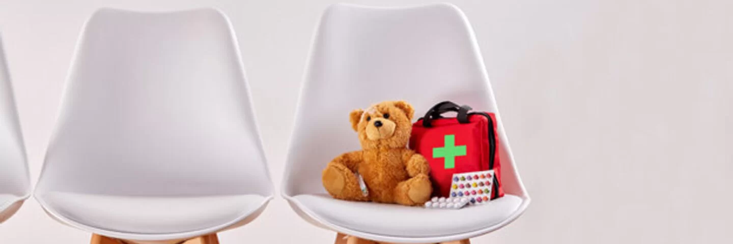 Das Bild zeigt vier weiße nebeneinander stehende Stühle. Auf dem Stuhl ganz rechts befindet sich ein Teddybär und ein kleiner Erste-Hilfe-Rucksack. 