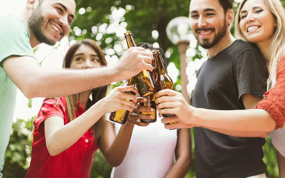 Fünf junge Leute stoßen im Garten mit Bierflaschen an. Die Gesichter sind angeschnitten und nicht zu erkennen.