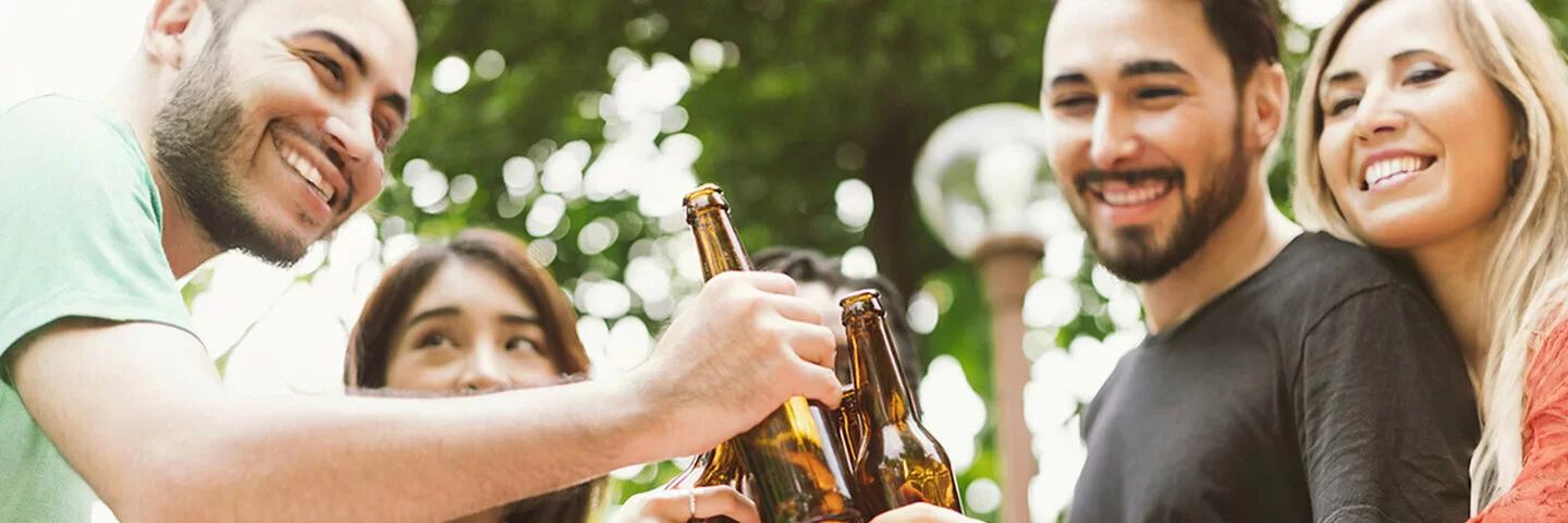 Fünf junge Leute stoßen im Garten mit Bierflaschen an. Die Gesichter sind angeschnitten und nicht zu erkennen.