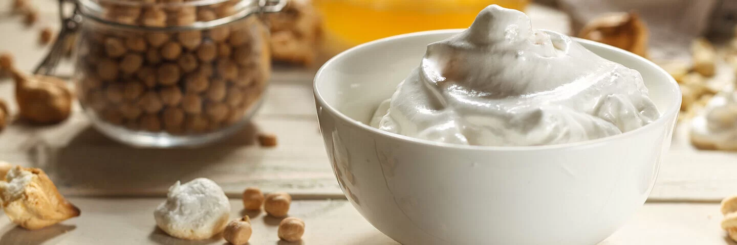 Auf einem Holztisch stehen ein Einmachglas Kichererbsen, eine Schüssel mit Abtropfwasser und aufgeschlagenes Aquafaba.