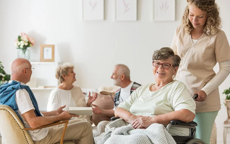 Eine Seniorin sitzt im Rollstuhl und wird von einer Pflegefachfrau geschoben. Im Hintergrund sind zwei Männer und eine Frau zu sehen, die sich unterhalten.